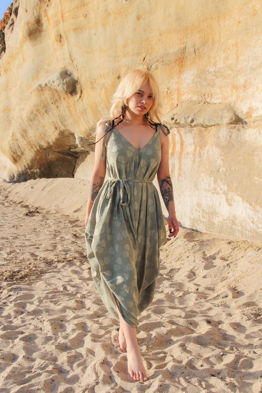 A person with blonde hair walks barefoot on a sandy beach, wearing a flowing green dress that resembles the elegance of a long jumpsuit. The backdrop features a large rock formation with a light brown and beige texture.