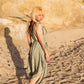 A woman with blonde hair and tattoos walks barefoot on a sunlit beach, gracefully clad in a light green long jumpsuit. Her shadow is cast on the sandy surface, and rock formations appear in the background.