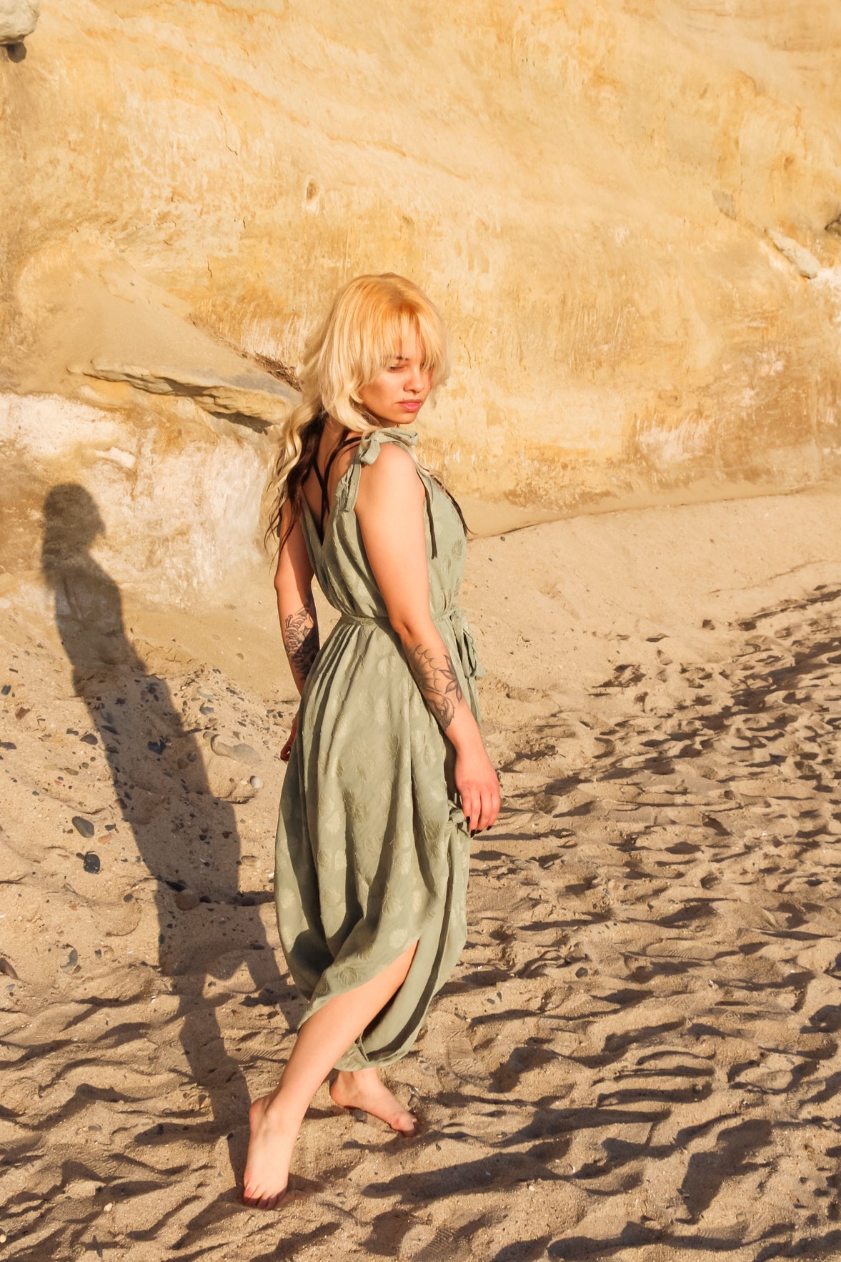 A woman with blonde hair and tattoos walks barefoot on a sunlit beach, gracefully clad in a light green long jumpsuit. Her shadow is cast on the sandy surface, and rock formations appear in the background.
