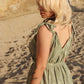 A person with blonde hair stands on a sandy beach, facing away. They wear a green, patterned long jumpsuit with tied shoulder straps. The outfit has a V-shaped back. A tattoo of leaves is visible on their arm. Rock formations are in the background. A person with blonde hair stands on a sandy beach, facing away. They wear a green, patterned long jumpsuit with tied shoulder straps. The outfit has a V-shaped back. A tattoo of leaves is visible on their arm. Rock formations are in the background.