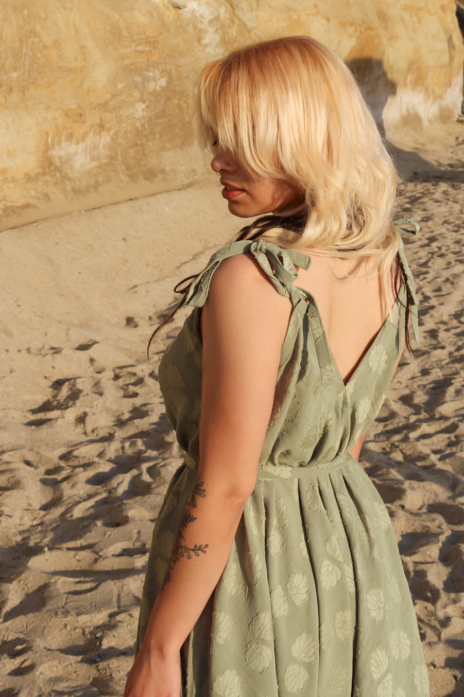 A person with blonde hair stands on a sandy beach, facing away. They wear a green, patterned long jumpsuit with tied shoulder straps. The outfit has a V-shaped back. A tattoo of leaves is visible on their arm. Rock formations are in the background. A person with blonde hair stands on a sandy beach, facing away. They wear a green, patterned long jumpsuit with tied shoulder straps. The outfit has a V-shaped back. A tattoo of leaves is visible on their arm. Rock formations are in the background.