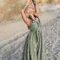 A woman with tattoos and long blonde hair poses gracefully on a sandy beach. She dons a flowing green dress resembling a long jumpsuit and is barefoot, with her arms extended upwards against a backdrop of rocky cliffs.