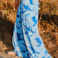 A person strolls on a dirt path, their flowing blue and white skirt with a palm leaf pattern resembling sexy pants with flowing panels. The skirt and part of an arm sway gracefully against the backdrop of dry grass and foliage.