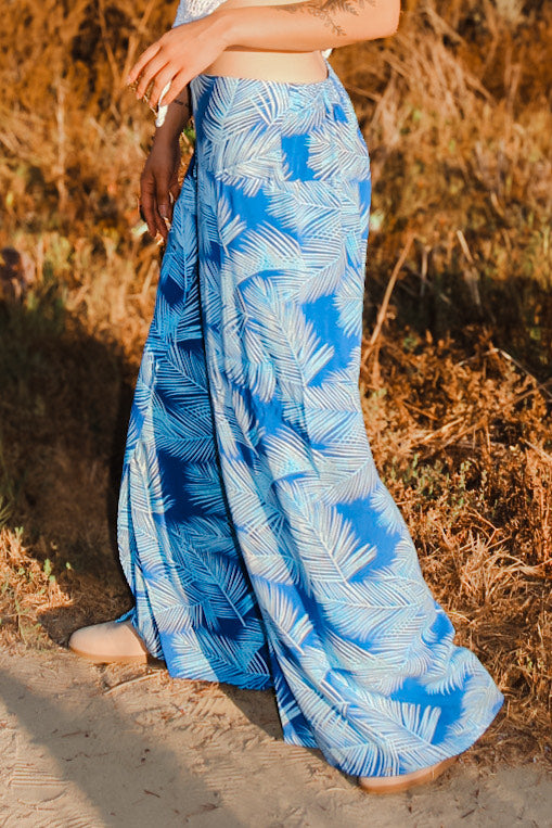 A person strolls on a dirt path, their flowing blue and white skirt with a palm leaf pattern resembling sexy pants with flowing panels. The skirt and part of an arm sway gracefully against the backdrop of dry grass and foliage.