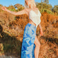 A person with blonde hair poses outdoors on a sunny day, wearing a white crocheted top and sexy pants with flowing panels in a blue leaf pattern. They stand on a path surrounded by trees and shrubs, casting a slight shadow on the ground.