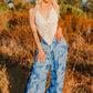A person with long blonde hair is wearing a white crochet halter top and sexy pants with flowing panels adorned in a blue tropical pattern, standing outdoors with dry foliage and trees in the background. The warm lighting hints at either sunset or sunrise.