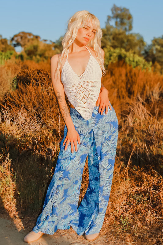 A person with long blonde hair is wearing a white crochet halter top and sexy pants with flowing panels adorned in a blue tropical pattern, standing outdoors with dry foliage and trees in the background. The warm lighting hints at either sunset or sunrise.