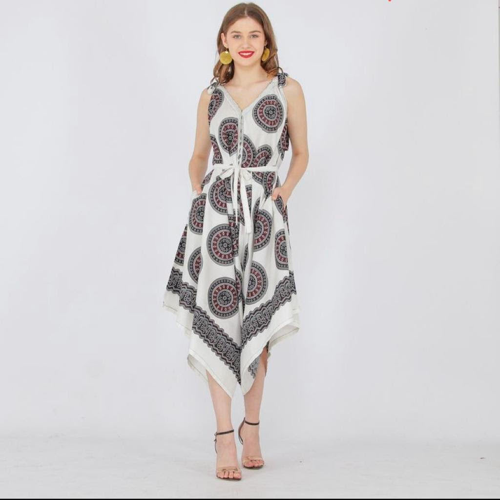 The woman dons a sleeveless dress with a geometric pattern, reminiscent of a chic long jumpsuit, standing against a white background. The V-neck and asymmetric hem are complemented by strappy heels and hoop earrings, while her hands rest casually in the dress pockets.