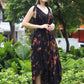 A woman stands outdoors on grass in a black and orange tie-dye long jumpsuit, gracefully holding the fabric at her waist. Lush green plants and trees form the backdrop, with red and white structures partially visible.