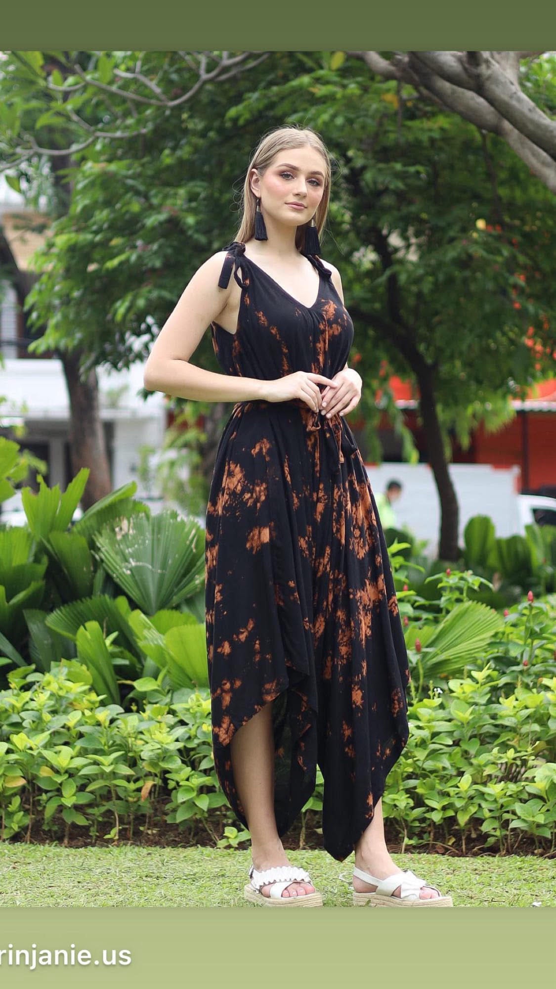 A woman stands outdoors on grass in a black and orange tie-dye long jumpsuit, gracefully holding the fabric at her waist. Lush green plants and trees form the backdrop, with red and white structures partially visible.