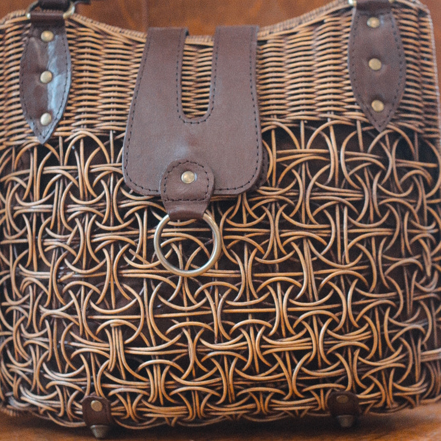 A large rattan bag with intricate patterns and leather accents. The front features a leather flap with a metal ring detail, blending rustic texture with modern elements for a striking look.