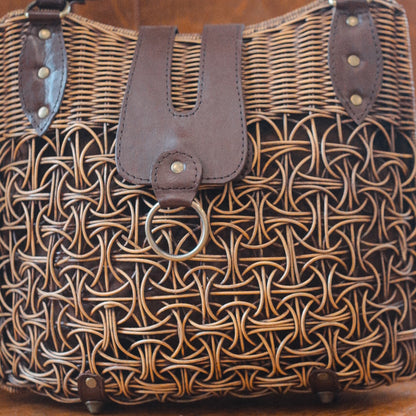 A large rattan bag with intricate patterns and leather accents. The front features a leather flap with a metal ring detail, blending rustic texture with modern elements for a striking look.