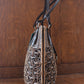 A stylish woven rattan purse with dark leather handles sits on a wooden surface. The intricate design features a mix of tightly woven and looser circular patterns, creating a textured look. The background is a wood panel surface.