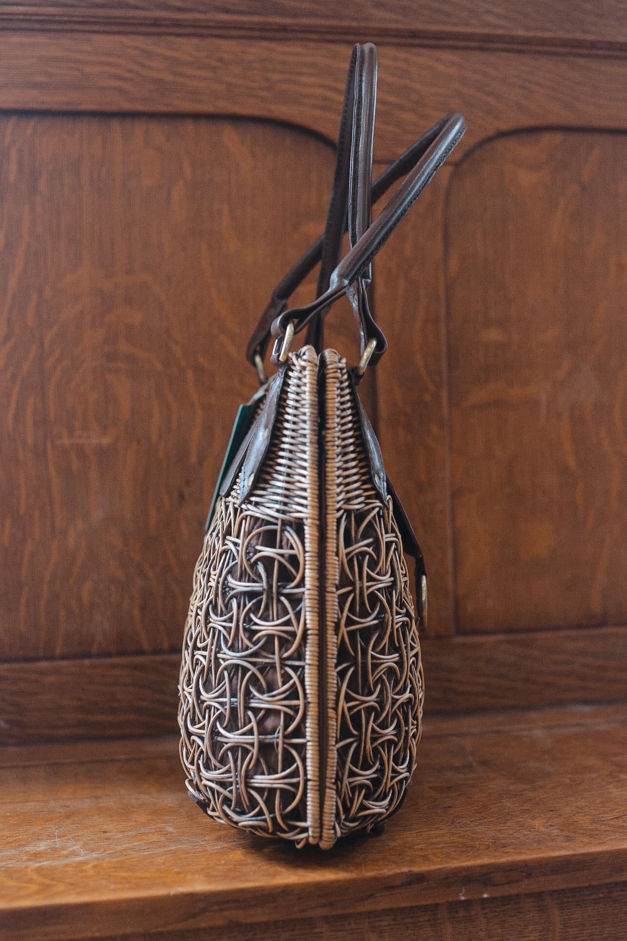 A stylish woven rattan purse with dark leather handles sits on a wooden surface. The intricate design features a mix of tightly woven and looser circular patterns, creating a textured look. The background is a wood panel surface.
