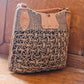 A large woven rattan purse with intricate designs and leather accents sits on a wooden surface. The bags pattern consists of interlocking circular shapes, and it features metal rivets for added detail.