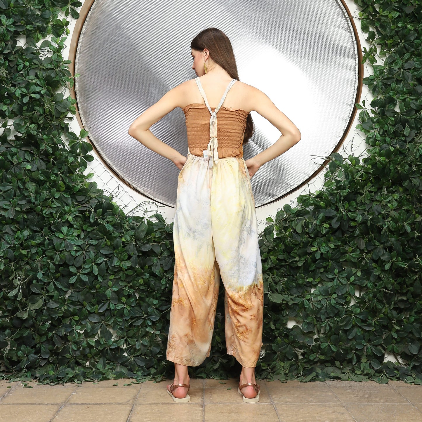 A woman stands with her back facing the camera, wearing colorful wide-leg overalls. She is in front of a circular metallic background, framed by lush green foliage.