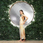 Woman in a tie dye overalls stands in front of a large circular mirror, framed by lush green leaves. She faces slightly to the side, with one hand on her waist. The setting has a natural, elegant ambiance.
