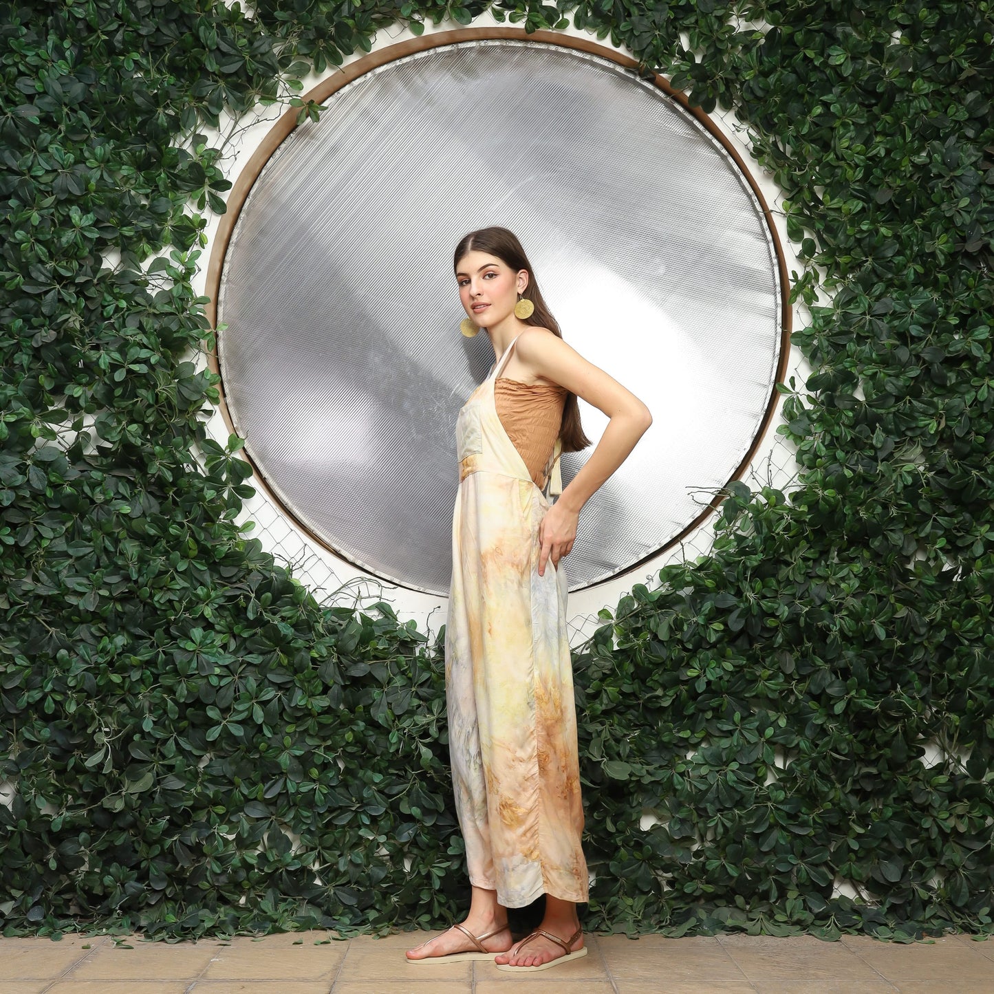Woman in a tie dye overalls stands in front of a large circular mirror, framed by lush green leaves. She faces slightly to the side, with one hand on her waist. The setting has a natural, elegant ambiance.