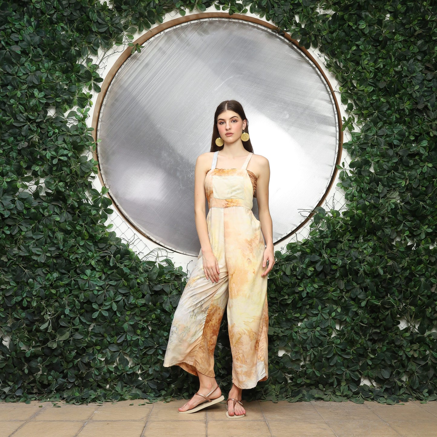 A woman in a flowy, earth-toned overalls stands in front of a large metallic circle, framed by lush green foliage. Sporting cute overalls for women, she has long brown hair and sandals, standing gracefully on a paved surface.