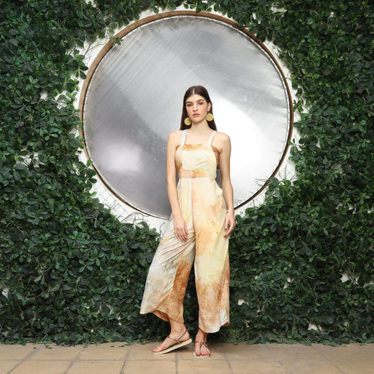 A woman in a flowy, earth-toned overalls stands in front of a large metallic circle, framed by lush green foliage. Sporting cute overalls for women, she has long brown hair and sandals, standing gracefully on a paved surface.