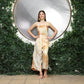 A woman stands confidently before a circular reflective surface surrounded by lush greenery. Dressed in beige and white tie-dye, she accessorizes with cute overalls for women layered stylishly and comfy sandals, her hands resting on her hips as she smiles warmly.