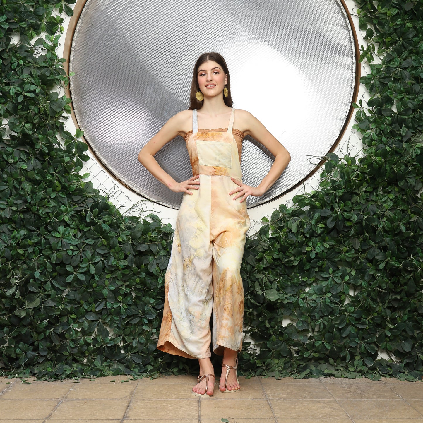 A woman stands confidently before a circular reflective surface surrounded by lush greenery. Dressed in beige and white tie-dye, she accessorizes with cute overalls for women layered stylishly and comfy sandals, her hands resting on her hips as she smiles warmly.
