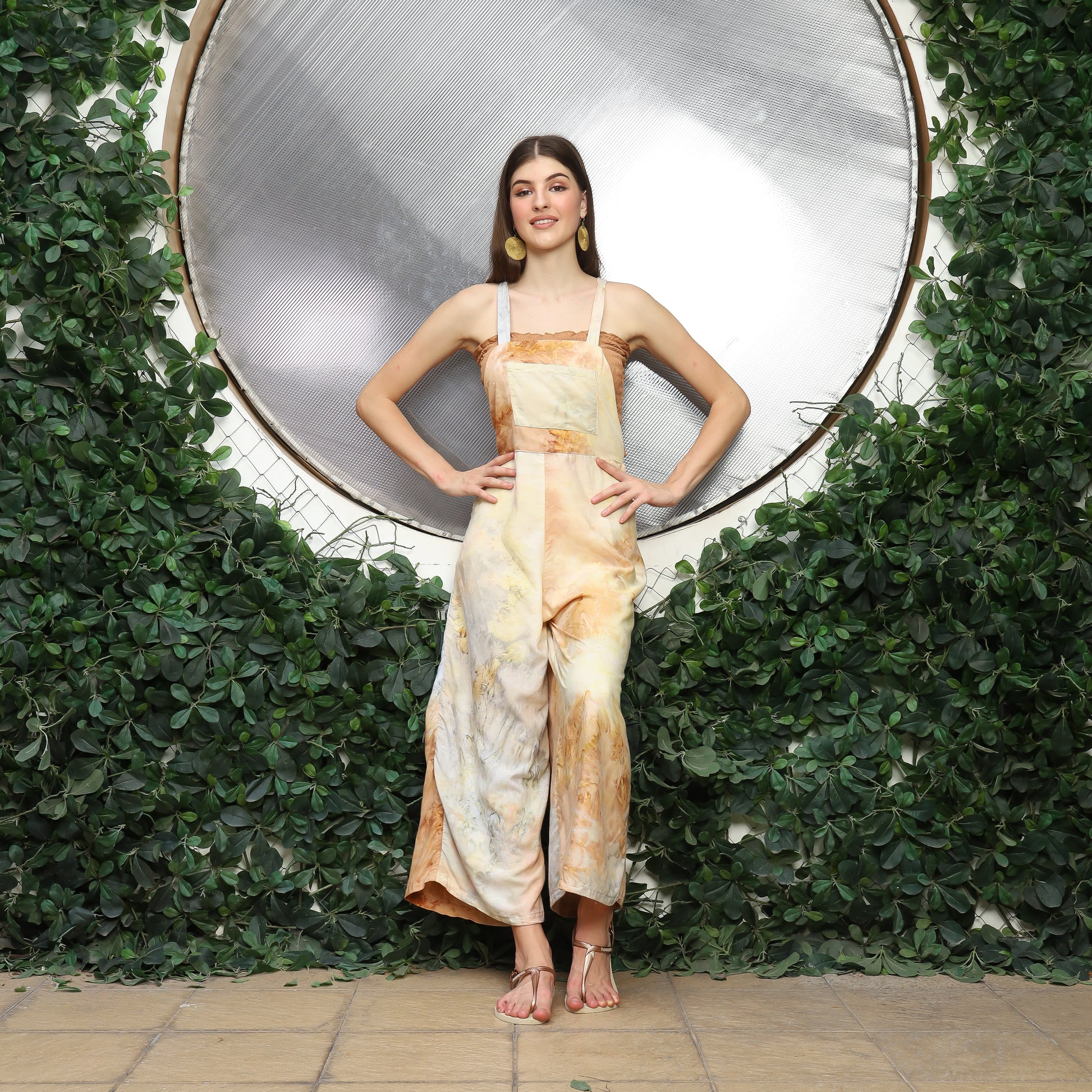 A woman stands confidently before a circular reflective surface surrounded by lush greenery. Dressed in beige and white tie-dye, she accessorizes with cute overalls for women layered stylishly and comfy sandals, her hands resting on her hips as she smiles warmly.