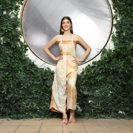 A woman stands confidently before a circular reflective surface surrounded by lush greenery. Dressed in beige and white tie-dye, she accessorizes with cute overalls for women layered stylishly and comfy sandals, her hands resting on her hips as she smiles warmly.