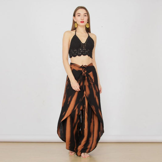 A woman poses against a white background wearing a black crochet halter top and tie-dye summer pants with flowing panels. She accessorizes with yellow hoop earrings and stands barefoot, with her arms by her sides. A woman poses against a white background wearing a black crochet halter top and tie-dye summer pants with flowing panels. She accessorizes with yellow hoop earrings and stands barefoot, with her arms by her sides.