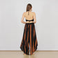 Woman in black halter top and flowing summer pants in orange-black tie-dye, standing barefoot on a wooden floor. Her back faces the camera, showcasing large round earrings. The scene has a light gray wall backdrop.
