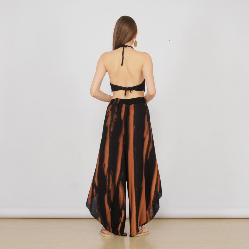 Woman in black halter top and flowing summer pants in orange-black tie-dye, standing barefoot on a wooden floor. Her back faces the camera, showcasing large round earrings. The scene has a light gray wall backdrop.