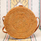 A round rattan purse with a woven design rests on a patterned textile backdrop, showcasing vertical stripes in beige, pink, yellow, and teal. The bag features a small loop closure and leather straps.