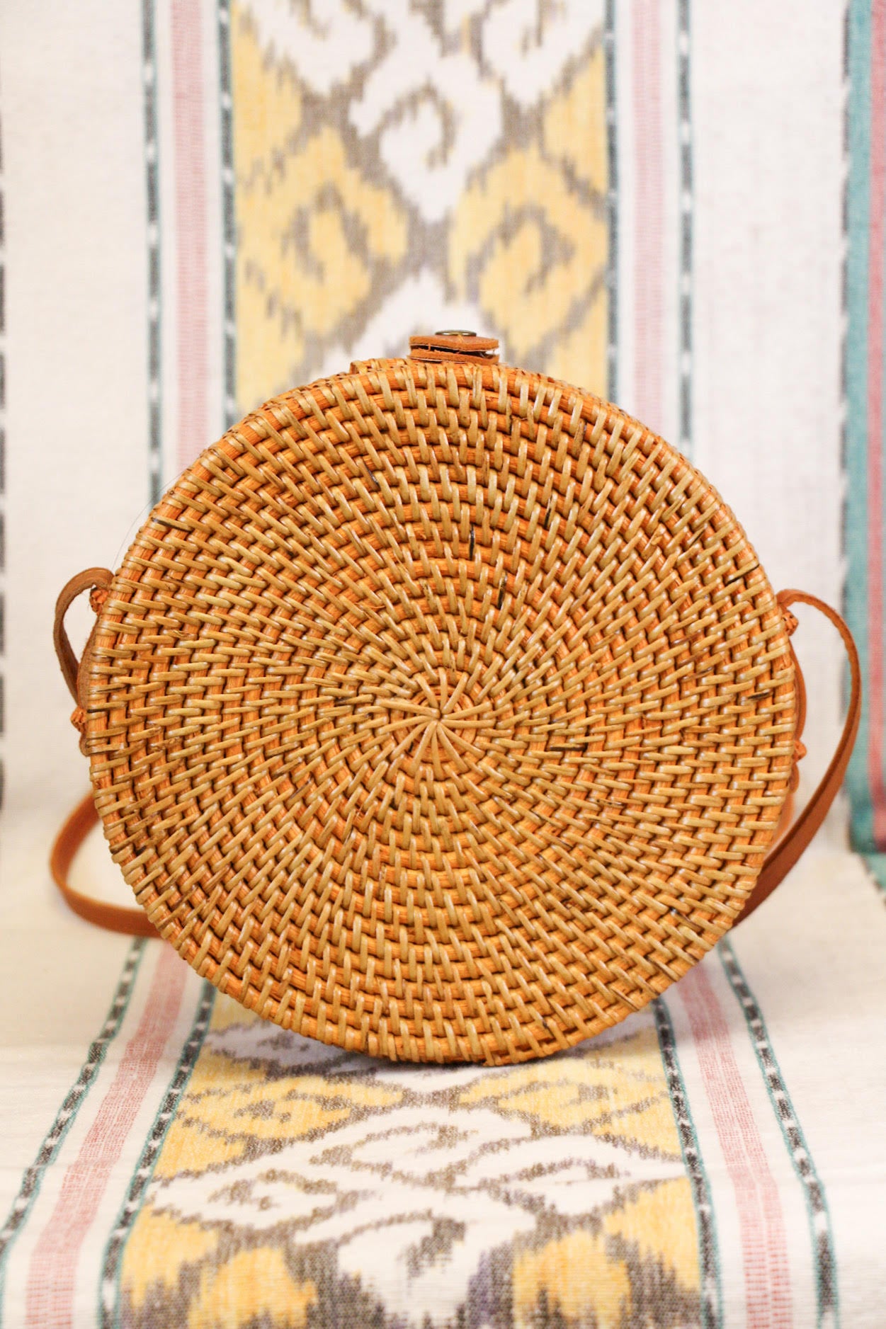 A round rattan purse with a woven design rests on a patterned textile backdrop, showcasing vertical stripes in beige, pink, yellow, and teal. The bag features a small loop closure and leather straps.