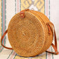 A round, woven rattan purse with a leather strap sits on a colorful patterned fabric background. The purse features intricate weaving and a simple design, highlighting its natural materials.