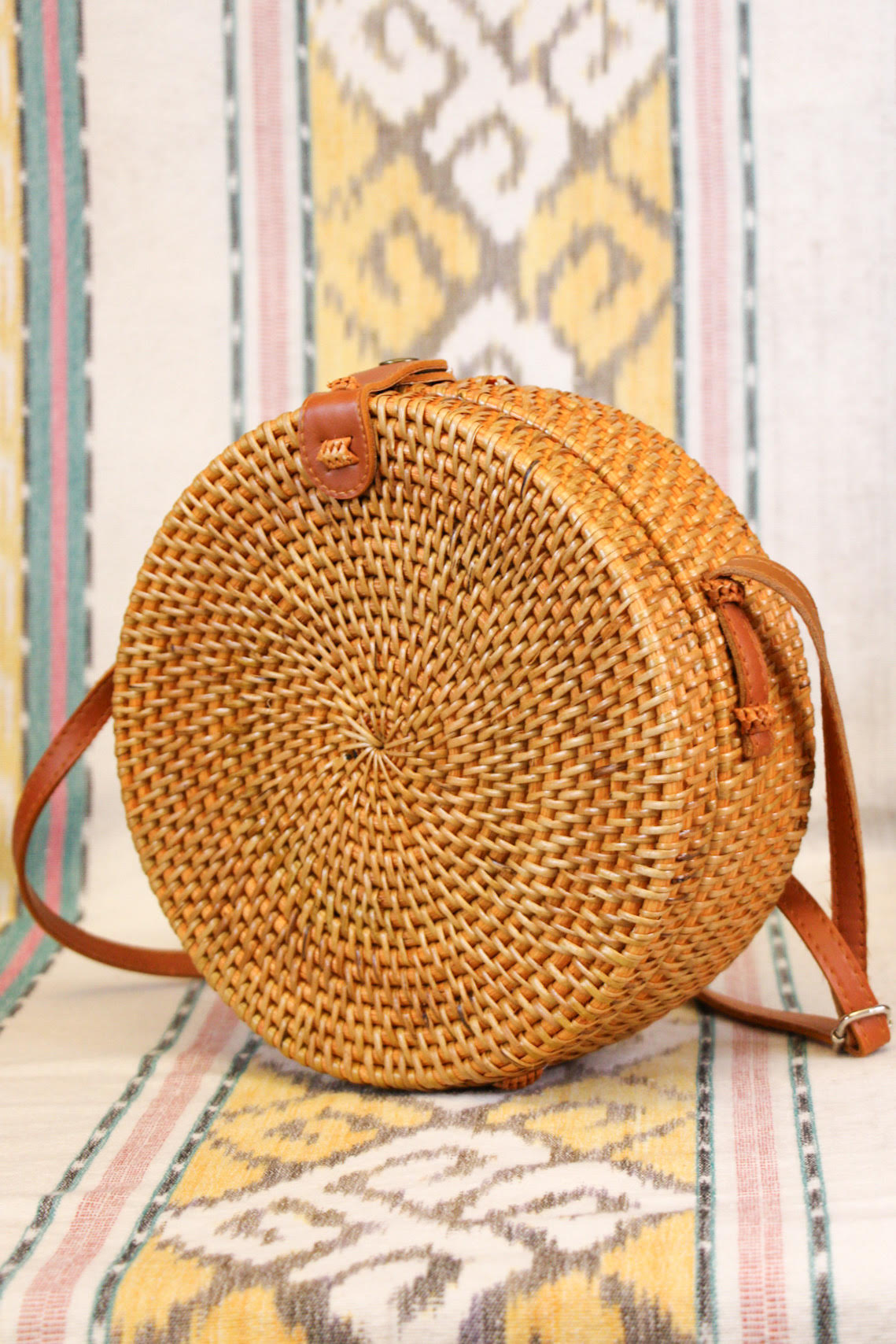 A round, woven rattan purse with a leather strap sits on a colorful patterned fabric background. The purse features intricate weaving and a simple design, highlighting its natural materials.