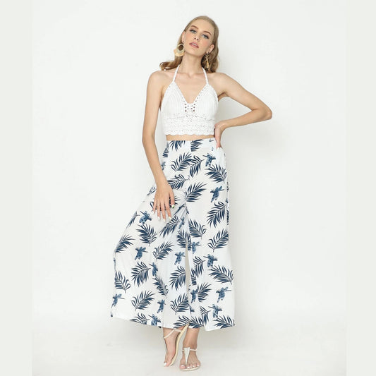 A woman in a white crop top and sexy pants with slits, adorned with a dark leaf pattern, poses against a light background. She wears hoop earrings and strappy heels, one hand on her hip while the other gently caresses her face.