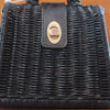A sleek black woven rattan purse with a leather strap and a brass twist-lock clasp on the front. The texture of the weave is beautifully visible, and the clasp is perfectly centered on the bag.