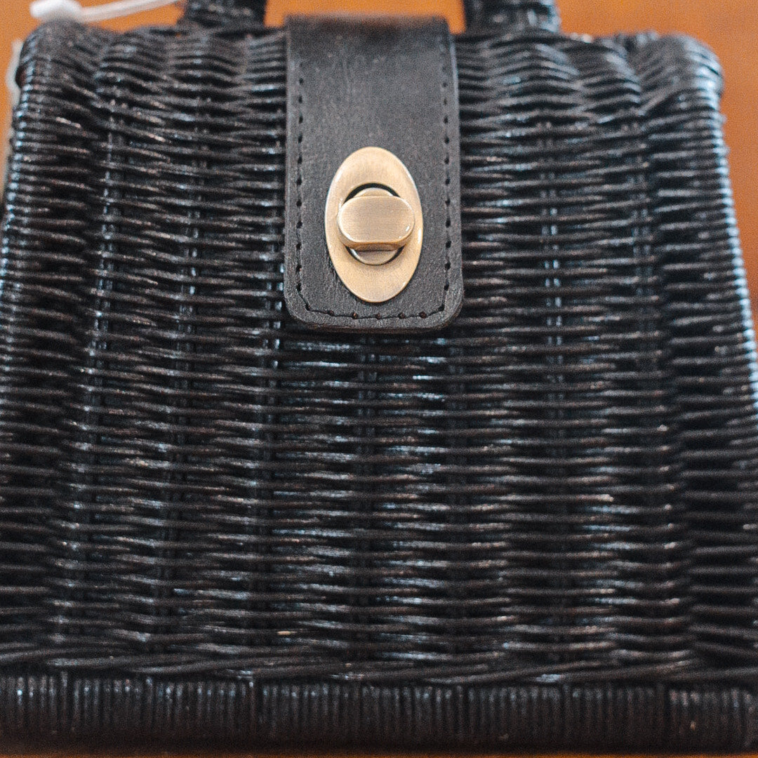 A sleek black woven rattan purse with a leather strap and a brass twist-lock clasp on the front. The texture of the weave is beautifully visible, and the clasp is perfectly centered on the bag.