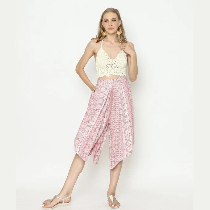 A woman poses against a white background, wearing a white crochet crop top and pink patterned high-waisted panel pants with diamond hem, paired with white sandals. Her long hair flows freely, and she completes the look with large hoop earrings.