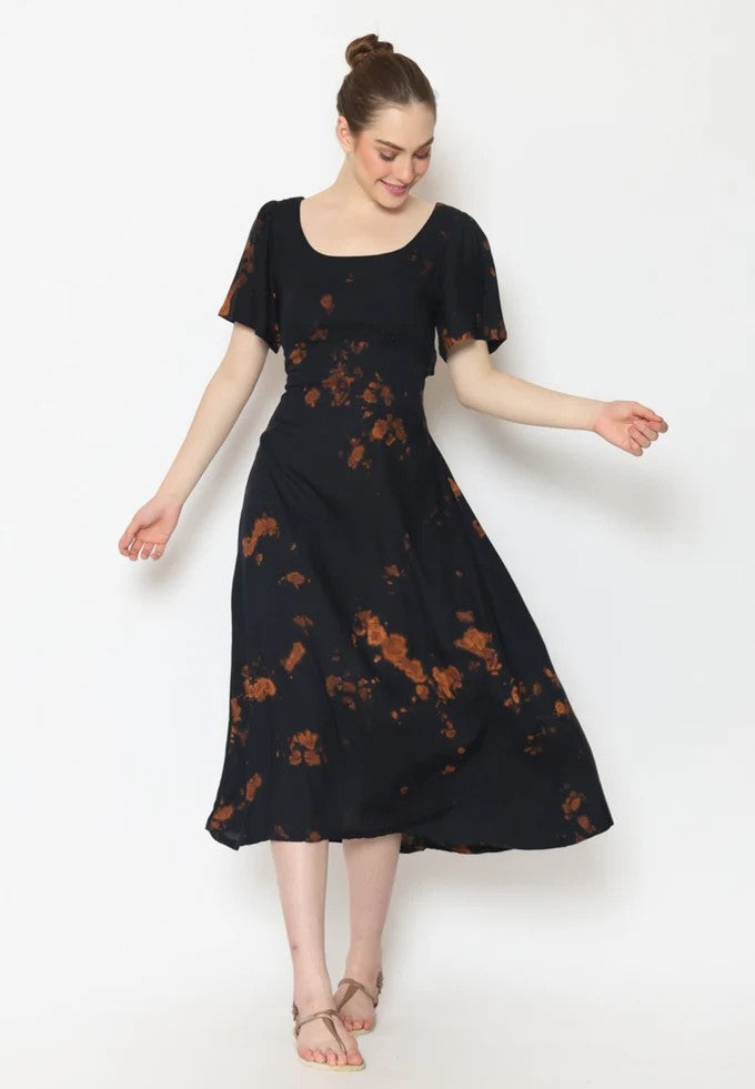 A woman poses in a dark, tie-dye patterned midi dress with short sleeves and a scoop neckline. Her hair is styled in a bun, reminiscent of the effortless charm of a tie back dress. She smiles, looking down with her arms slightly extended, barefoot against a white background.