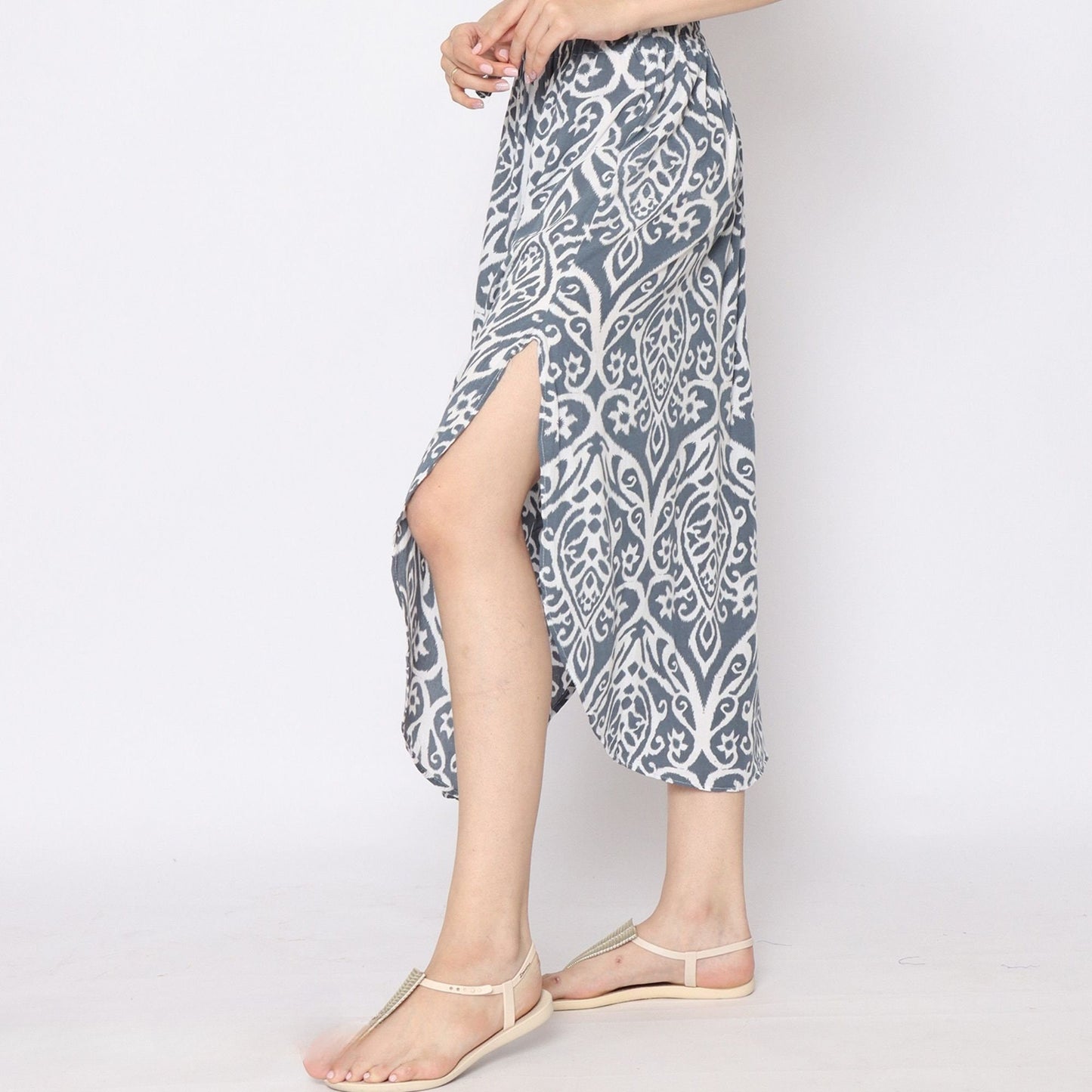 A person stands in profile wearing a blue and white skirt with side slits, showcasing its unique pattern. They pair it with beige flat sandals, posed elegantly with hands clasped near the waist. The backdrop is a simple, plain white.