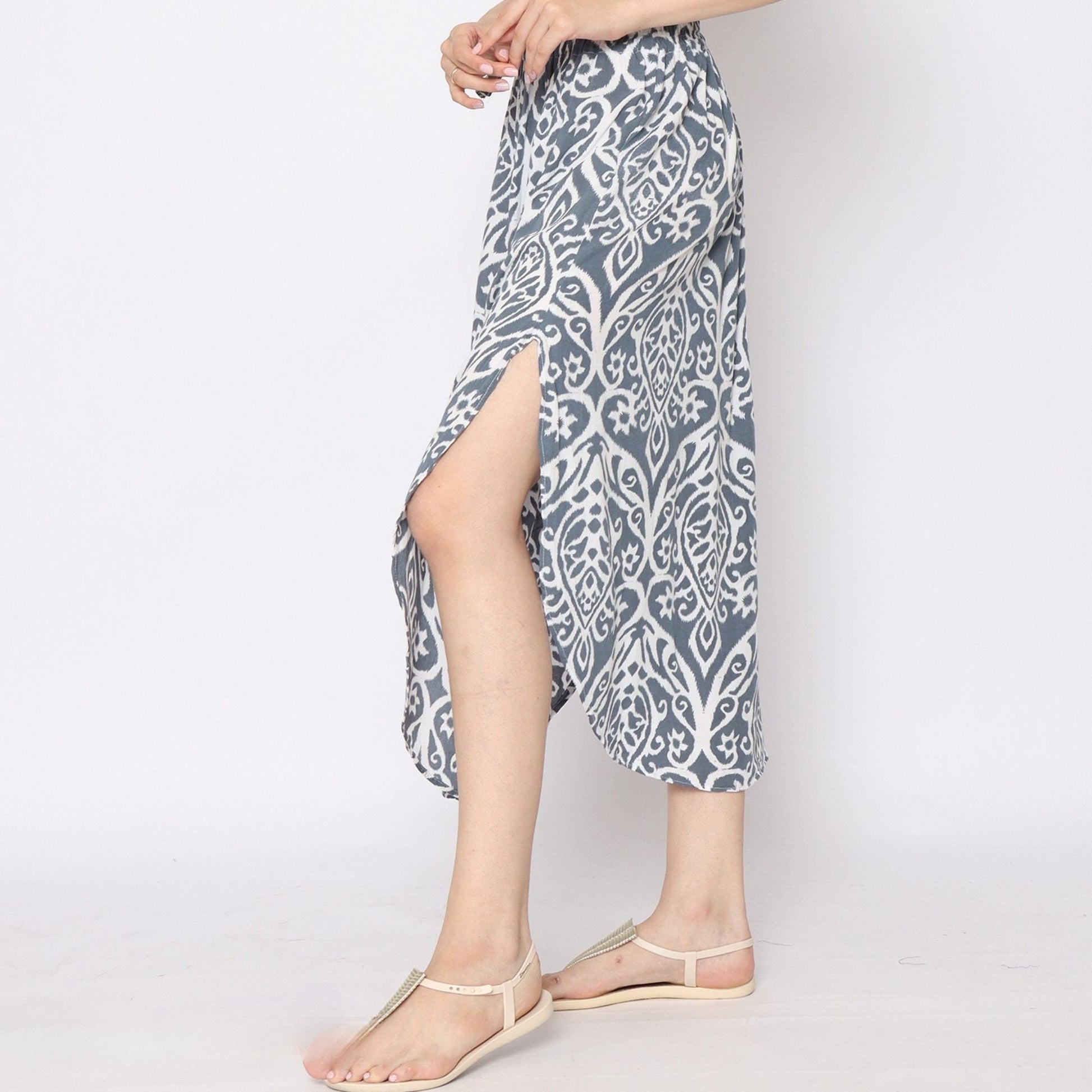 A person stands in profile wearing a blue and white skirt with side slits, showcasing its unique pattern. They pair it with beige flat sandals, posed elegantly with hands clasped near the waist. The backdrop is a simple, plain white.