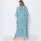 Woman in a blue and white floral-patterned caftan stands with her back to the camera against a plain white background. She has shoulder-length light brown hair and is wearing beige sandals.