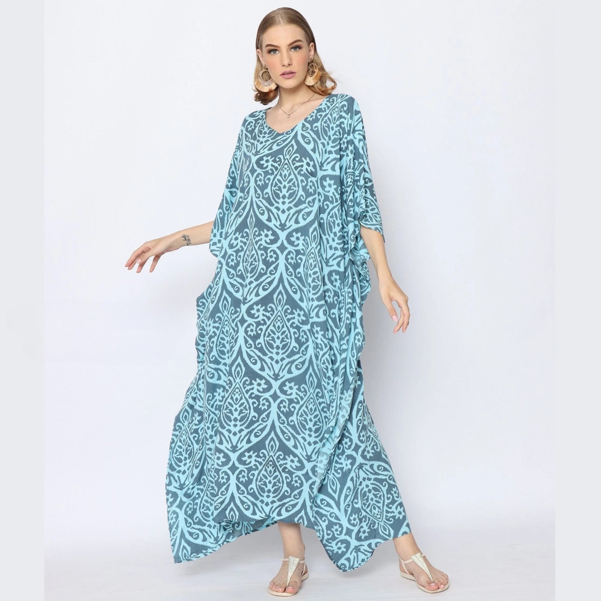 A woman in a flowing blue caftan with an intricate white pattern poses confidently. She stands against a plain background, wearing silver sandals and hoop earrings, her hair styled in waves.