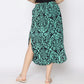 A person wearing a black top and a green and black patterned skirt with side slits, standing against a plain background. The skirt features an intricate floral design, and the person is wearing light-colored sandals.