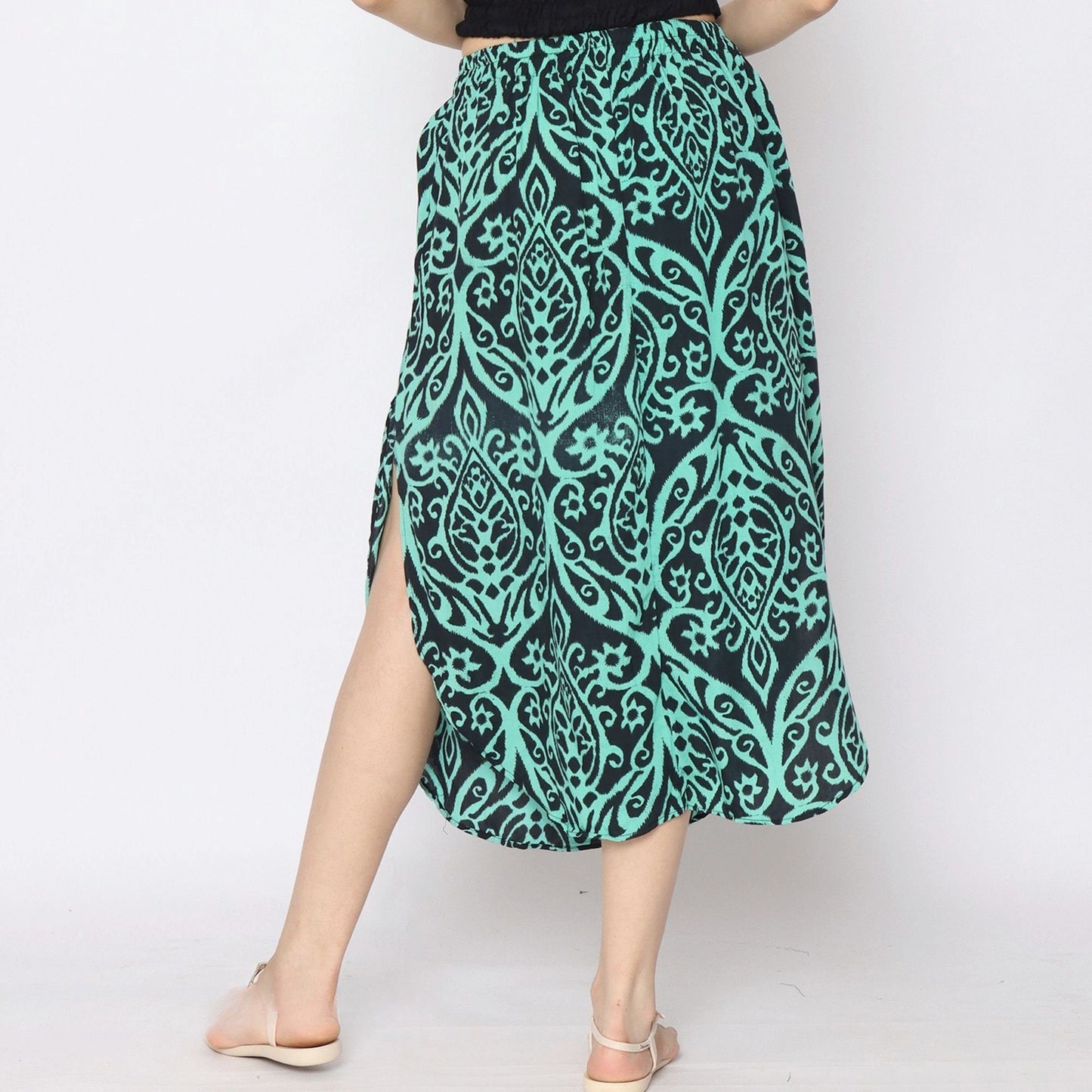 A person wearing a black top and a green and black patterned skirt with side slits, standing against a plain background. The skirt features an intricate floral design, and the person is wearing light-colored sandals.