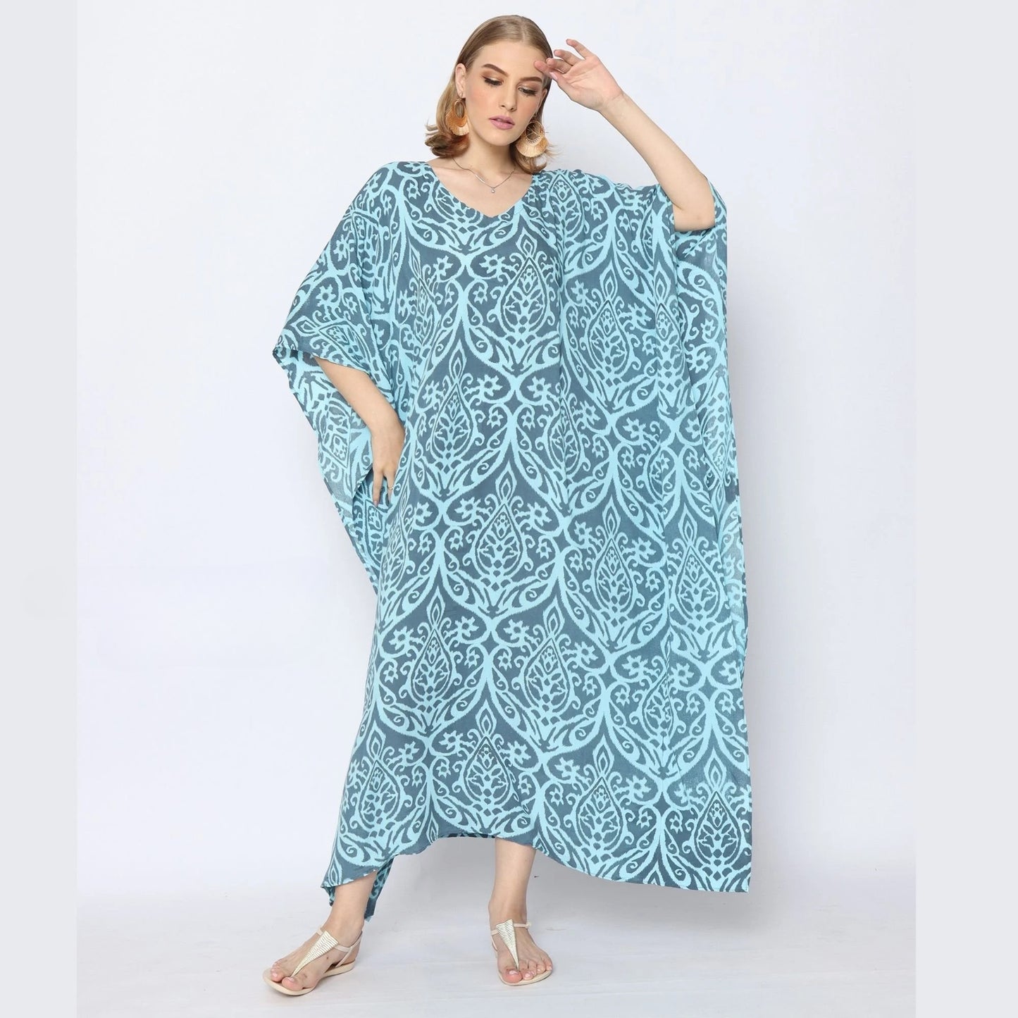 A woman in a flowing blue caftan with an intricate white pattern poses confidently. She stands against a plain background, wearing silver sandals and hoop earrings, her hair styled in waves.