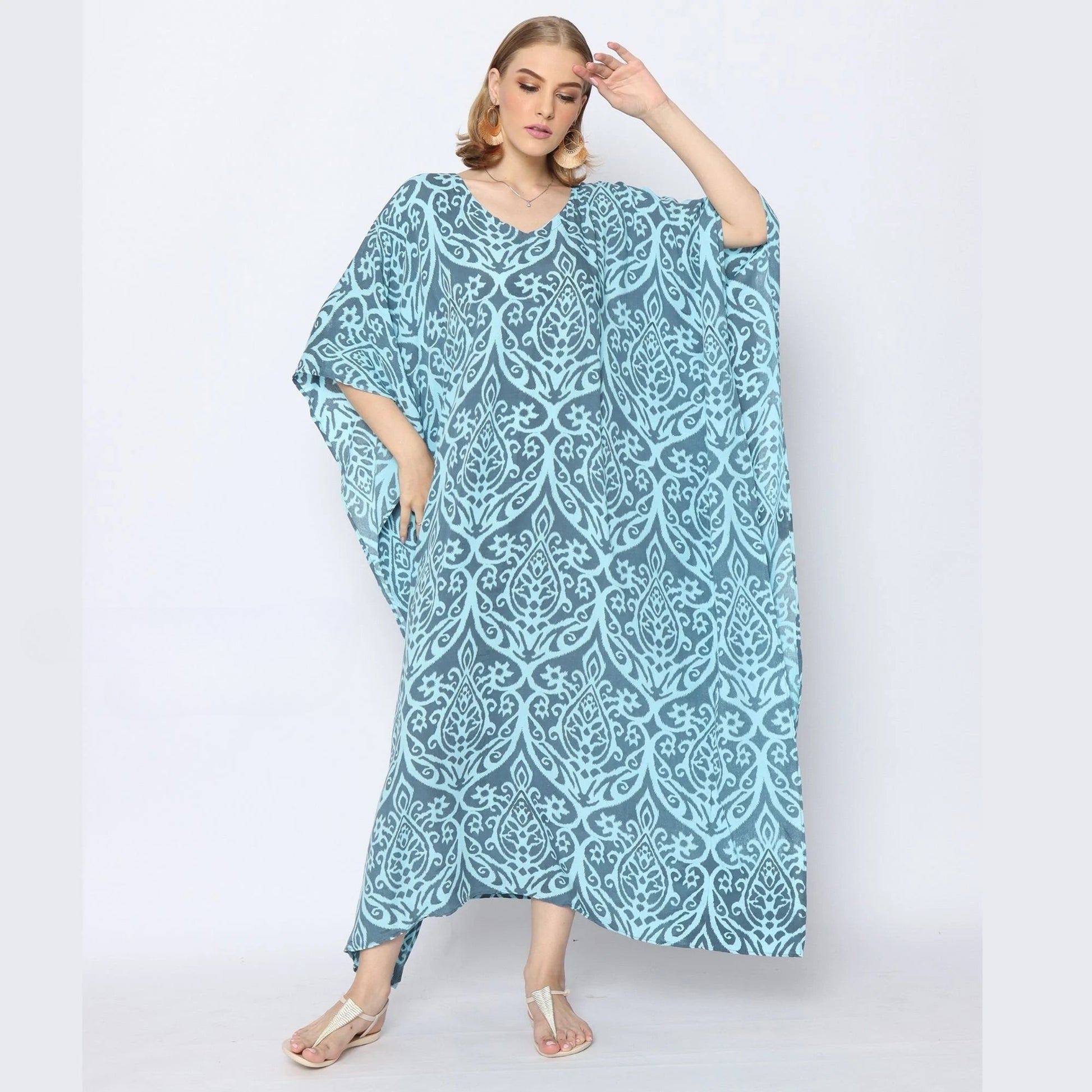 A woman in a flowing blue caftan with an intricate white pattern poses confidently. She stands against a plain background, wearing silver sandals and hoop earrings, her hair styled in waves.