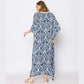 A woman is standing with her back to the camera, wearing a long, loose-fitting blue and white patterned dress. The caftan dress features an intricate white design on a dark blue background. She is wearing sandals and has shoulder-length brown hair.