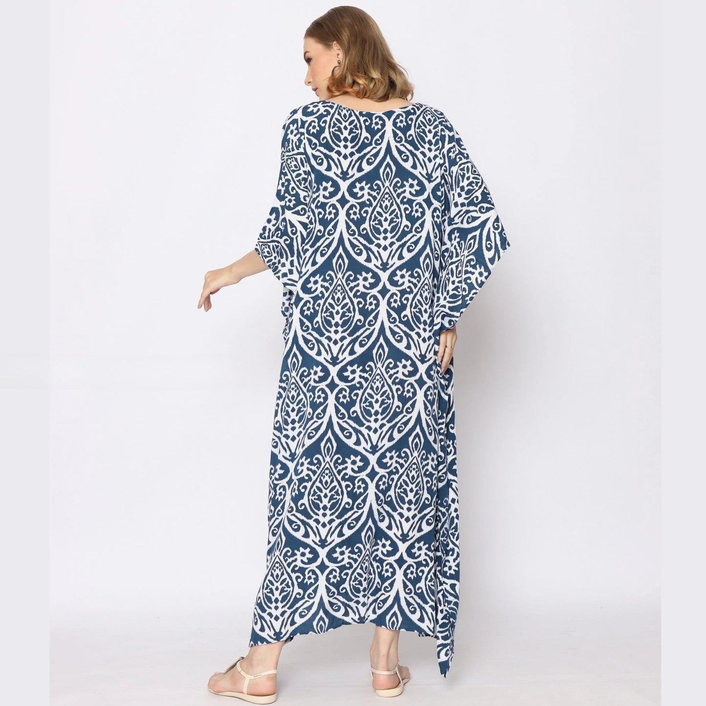 A woman is standing with her back to the camera, wearing a long, loose-fitting blue and white patterned dress. The caftan dress features an intricate white design on a dark blue background. She is wearing sandals and has shoulder-length brown hair.